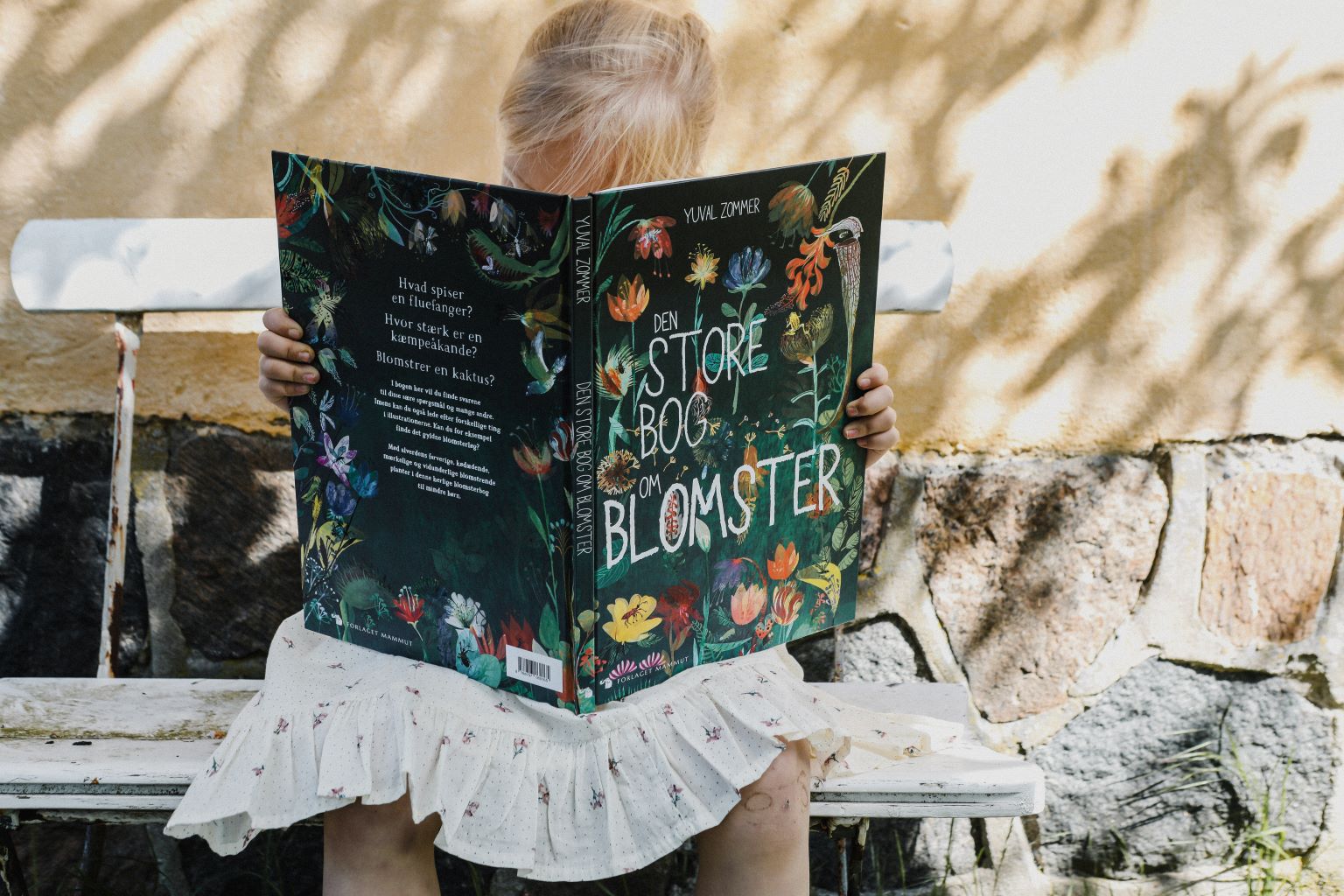 Barn læser "Den store bog om blomster" siddende udendørs på bænk