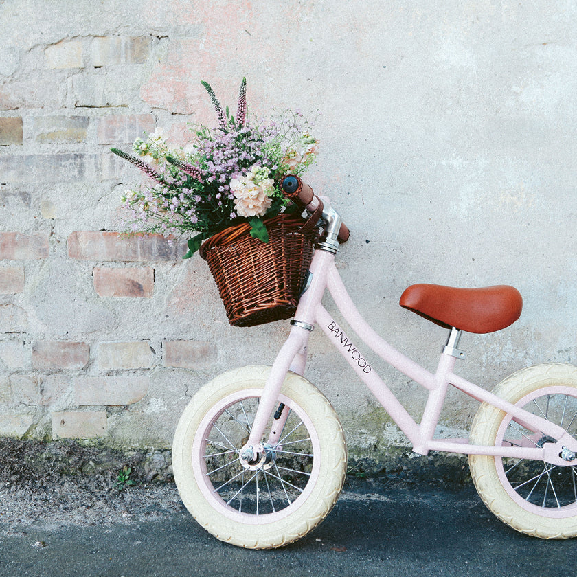 Lyserød Banwood børnecykel med fletkurv fyldt med blomster op ad mur